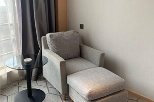Una habitación de esquina con un sillón gris, una mesa de vidrio y un objeto blanco, y una ventana con cortinas.