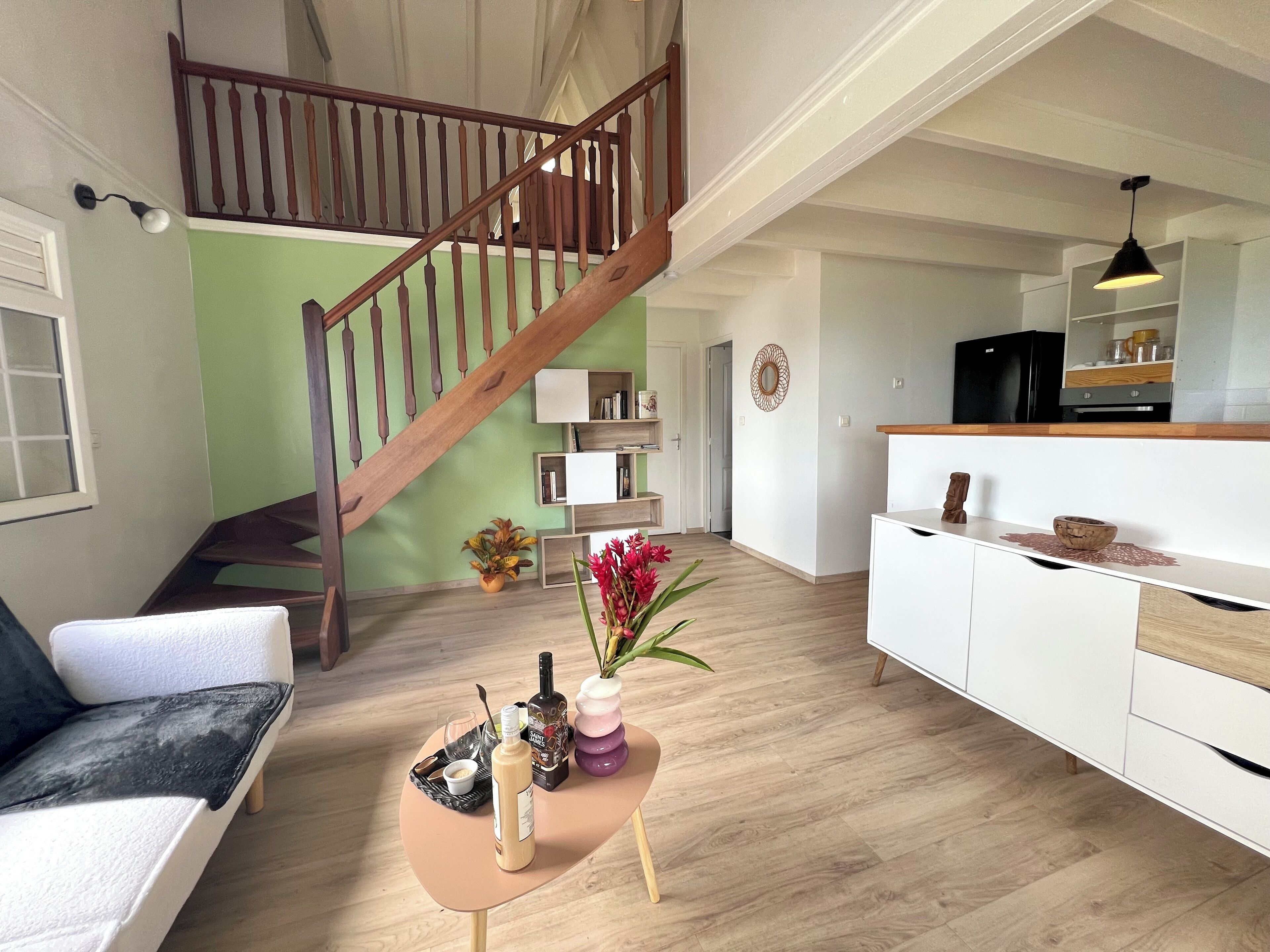 A spacious living area with a wooden staircase, a white sofa, a small round table with drinks, and a kitchen area with a black refrigerator.