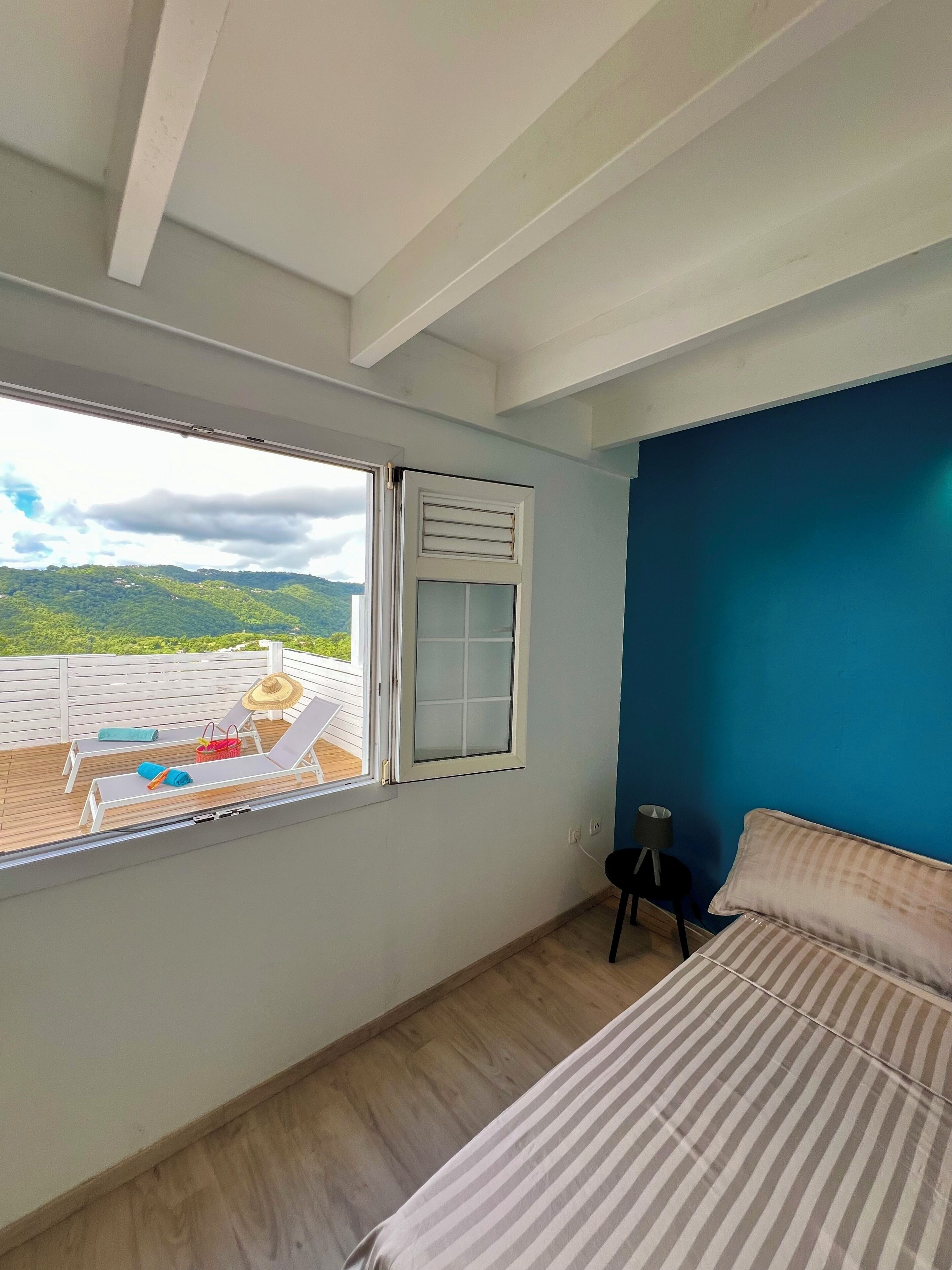 A bedroom with a bed, a window with a view of greenery, and a blue wall.
