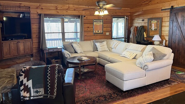L’intérieur chaleureux d’un chalet en bois comprend un canapé d’angle, un fauteuil en cuir, un plafond en bois et un ventilateur de plafond.