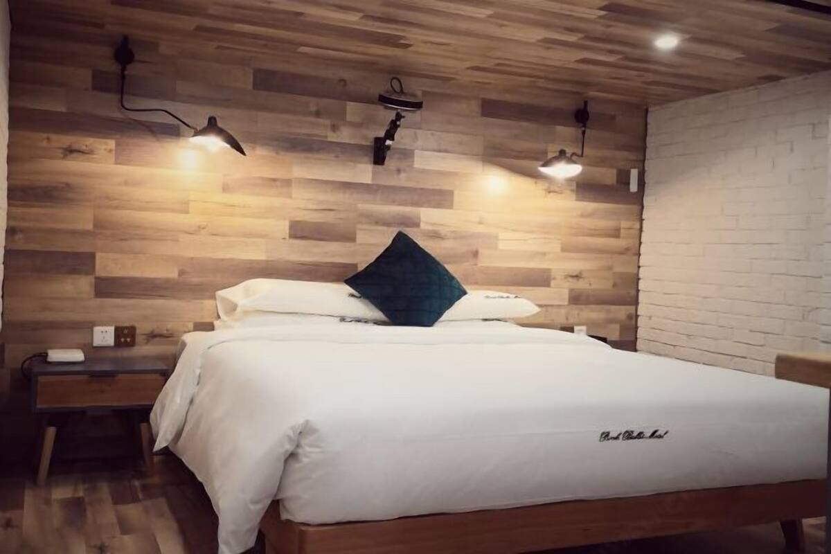 A neatly made bed with a white comforter and a dark pillow, a bedside table with a lamp, and a wooden wall with a brick accent.