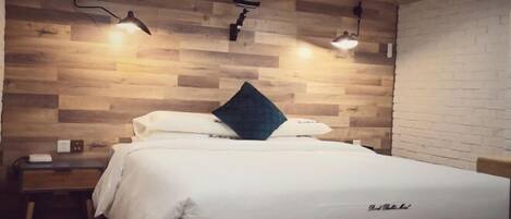Una cama bien hecha con un colchón blanco y un almohadón oscuro, una mesita de noche con una lámpara y una pared de madera con sección de ladrillo.