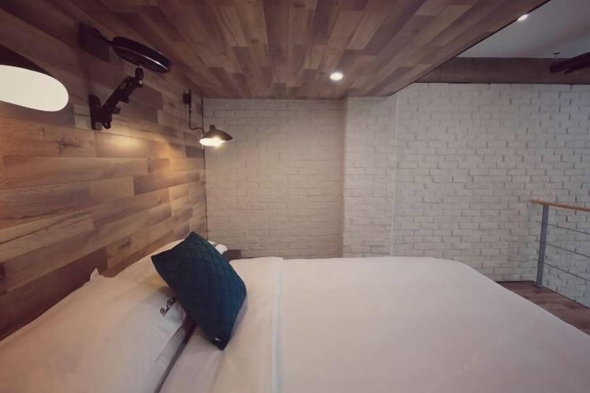 A bedroom with a wooden ceiling, a white brick wall, a bed with white bedding, and a nightstand with a lamp.