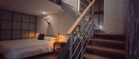 Un dormitorio con cama, mesita de noche, lámpara y una escalera con barandilla.