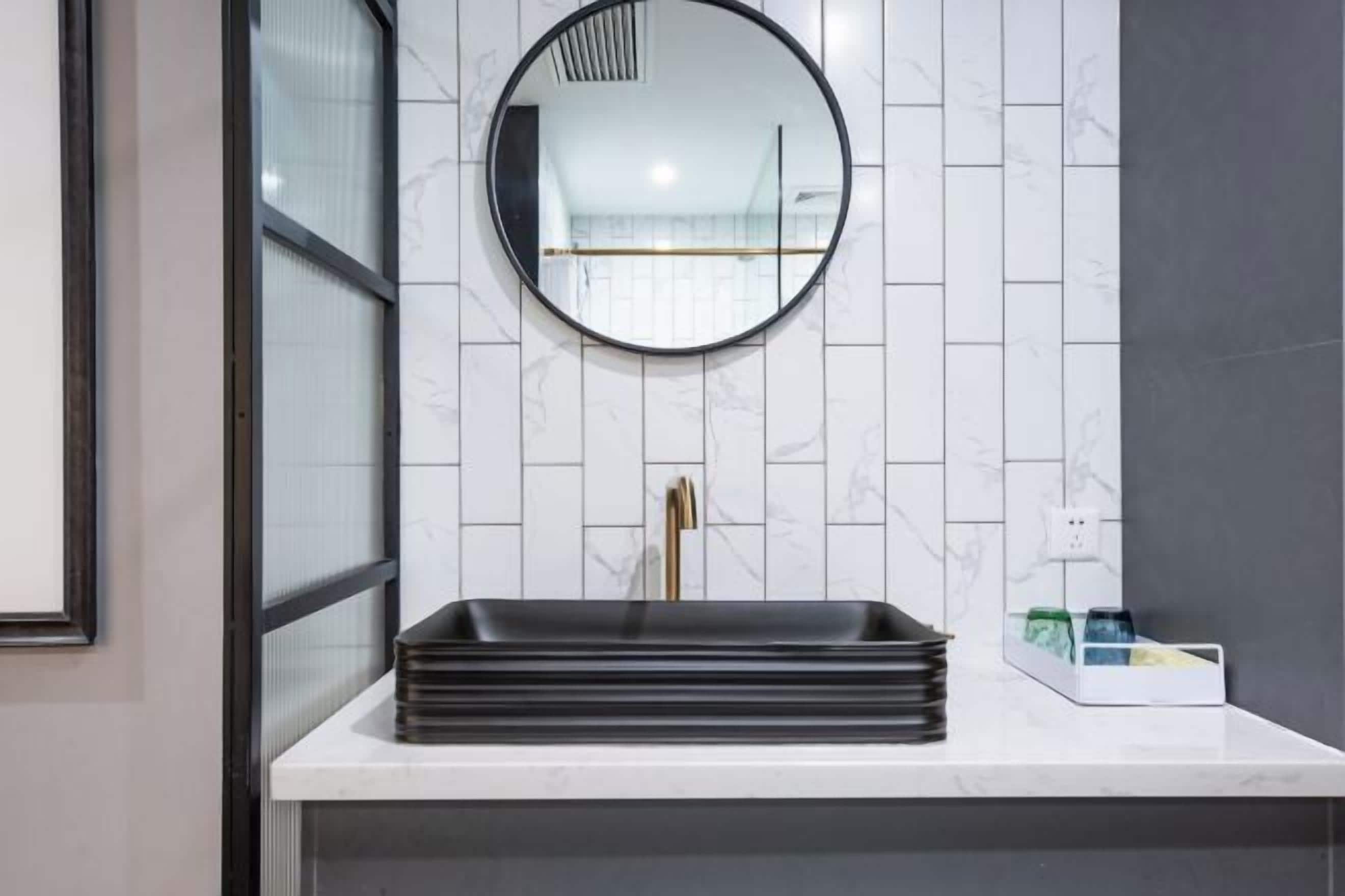 A bathroom with a white tiled wall, a round mirror, a black sink, and a gold faucet.
