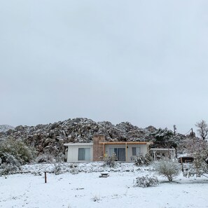 Ein modernes Haus mit flachem Dach, einer kleinen Veranda und einer Steinmauer im Hintergrund.