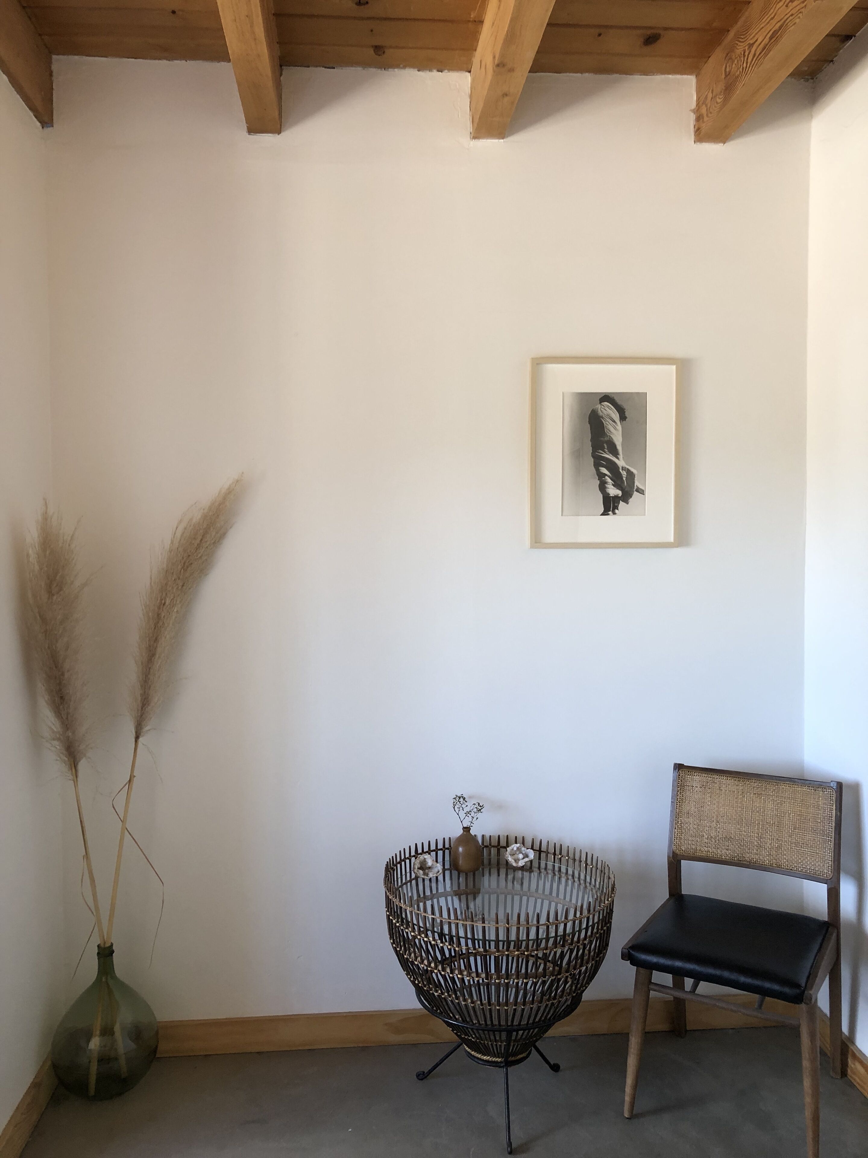 Una habitación con techo de madera, una fotografía blanco y negro enmarcada en la pared, una silla de mimbre, un banco negro acolchado y un jarrón con pastos secos.
