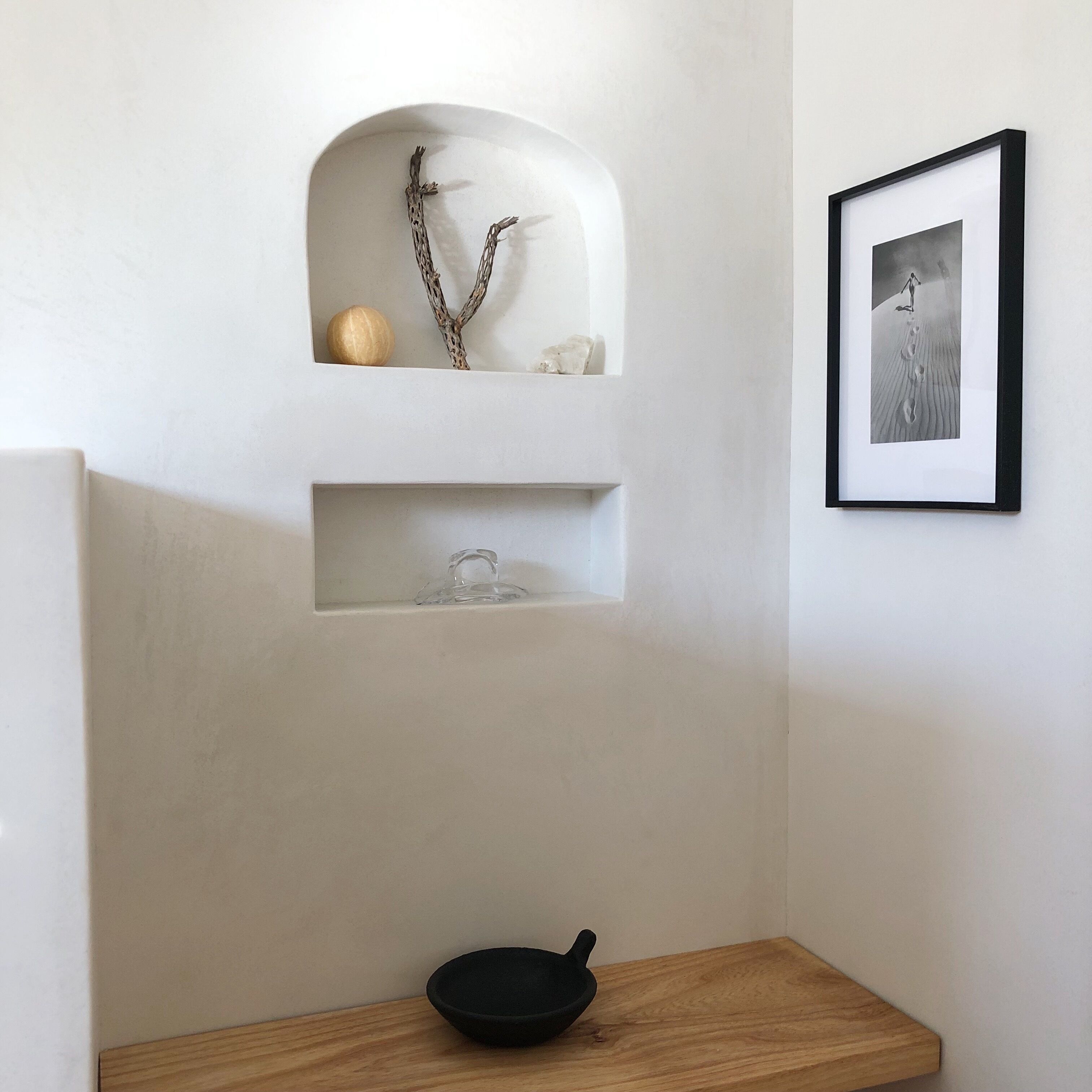 A minimalist room with a white wall, a wooden shelf with decorative items, a framed photograph, and a black bowl on a wooden table.