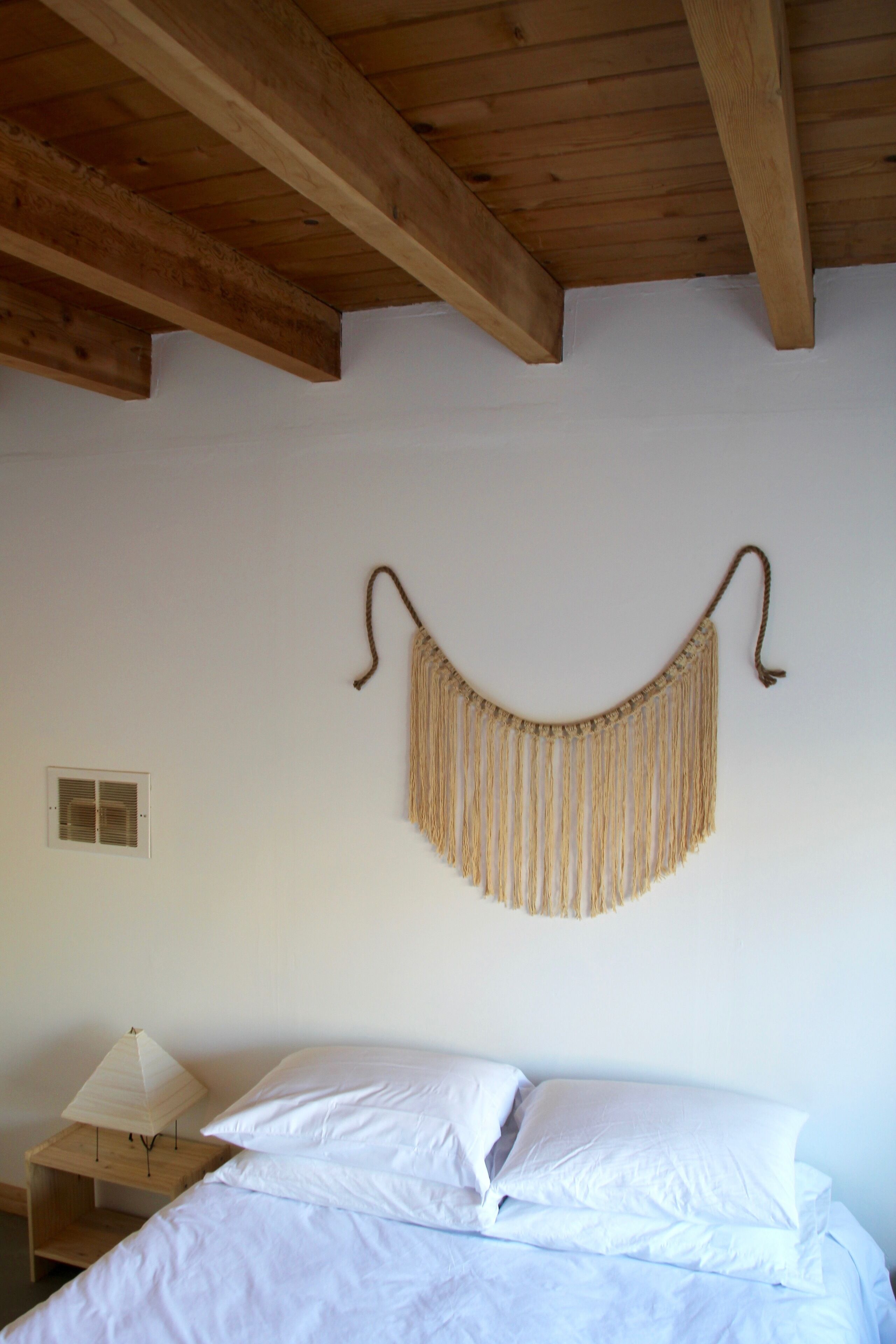 A bedroom with a bed, white bedding, a wooden nightstand with a lamp, and a wall-mounted macrame piece.