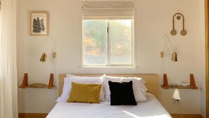 A bedroom with a wooden ceiling, a large bed with white bedding, two bedside tables with lamps, a window with blinds, and a wall-mounted picture. - Peaceful mid-century house under the boulders near Joshua Tree National Park (Joshua Tree)