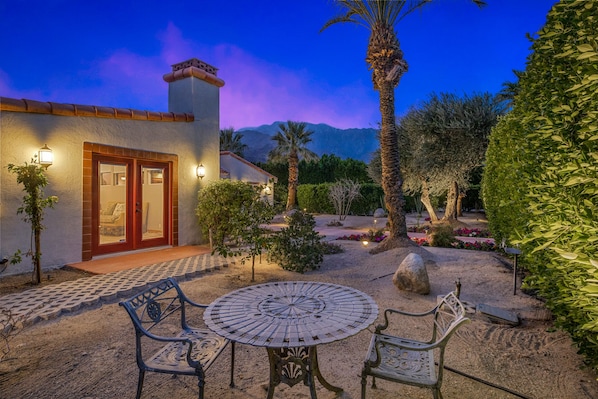 Un patio con una mesa y sillas redondas, un edificio con una puerta y luces fijadas a la pared, y un jardín con diversas plantas y una palmera.
