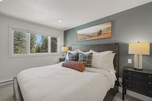 A bedroom with a large bed, a nightstand with a lamp, a window with a view of trees, and a painting on the wall.