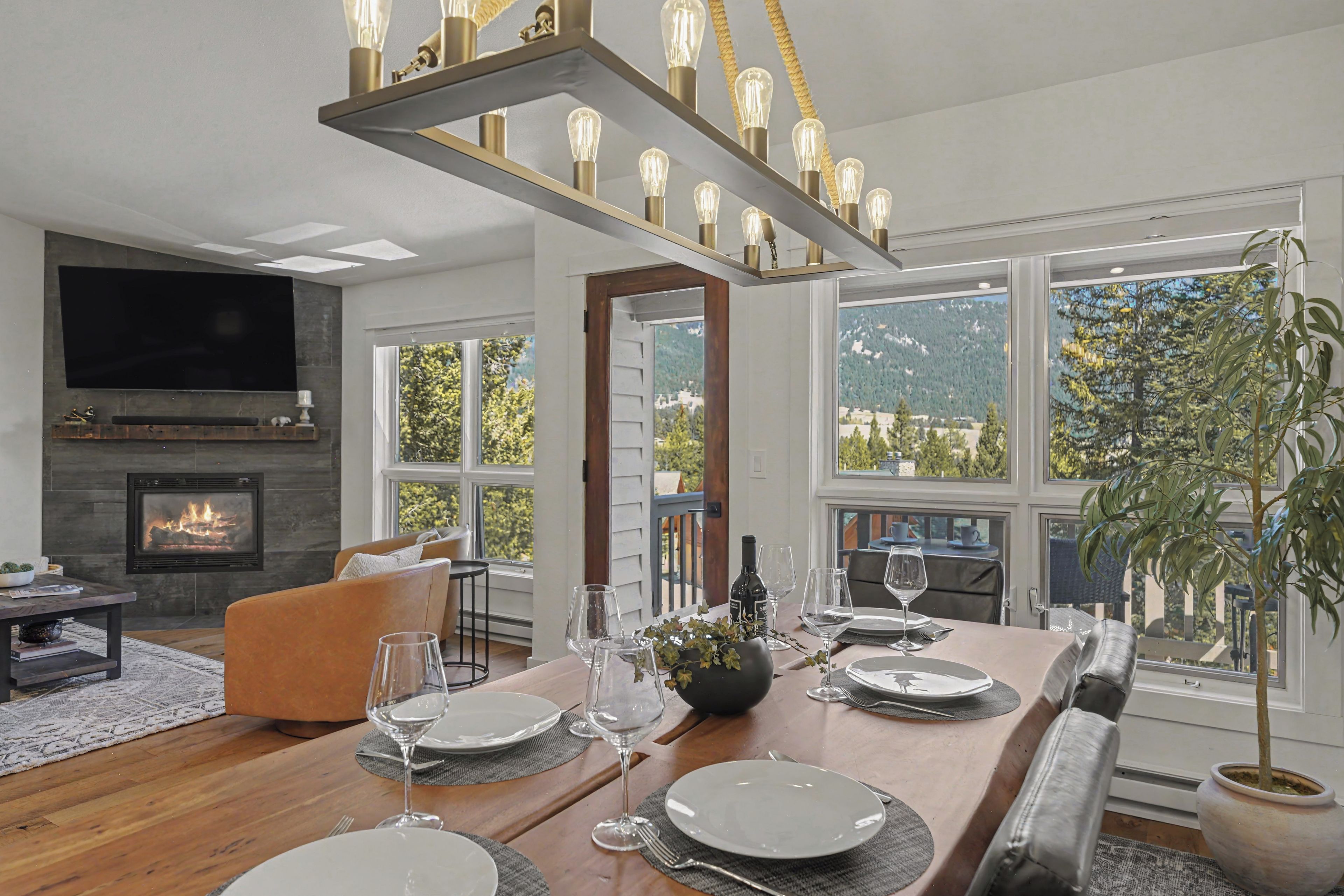 A modern living room with a fireplace, a dining area set for six, and large windows offering a view of the outdoors.