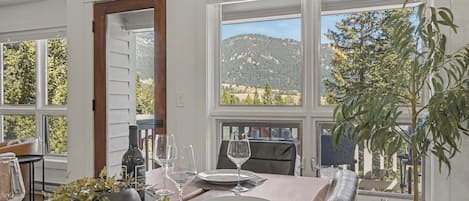 A dining area with a table set for two, a bottle of wine, and a view of a mountainous landscape through large windows.