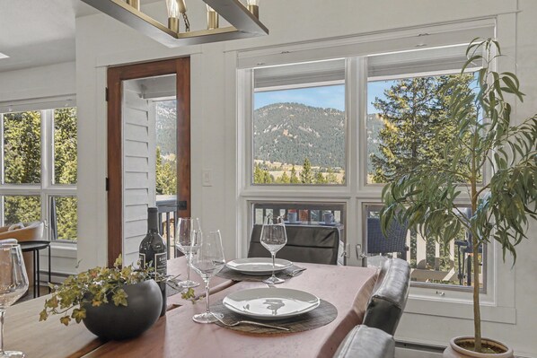 A dining area with a table set for two, a bottle of wine, and a view of a mountainous landscape through large windows.