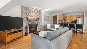 A modern living room with a stone fireplace, a flat-screen TV, a grey sofa, and a kitchen with wooden cabinets and stainless steel appliances.
