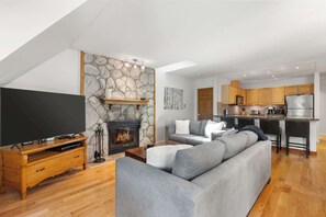 Ein modernes Wohnzimmer mit Kamin aus Stein, einem Flachbildfernseher, einem grauen Sofa und einer Küche mit Holzschränken sowie Edelstahlgeräten.