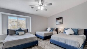 Ein Hotelzimmer mit drei Betten, einem Deckenventilator, einem großen Fenster und einem gerahmten Bild an der Wand.