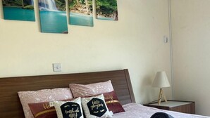 Cama bem arrumada com roupa de cama estampada, toalha enrolada e almofadas decorativas com texto. Há um ar condicionado fixado na parede e uma pequena mesa de cabeceira com uma luminária.
