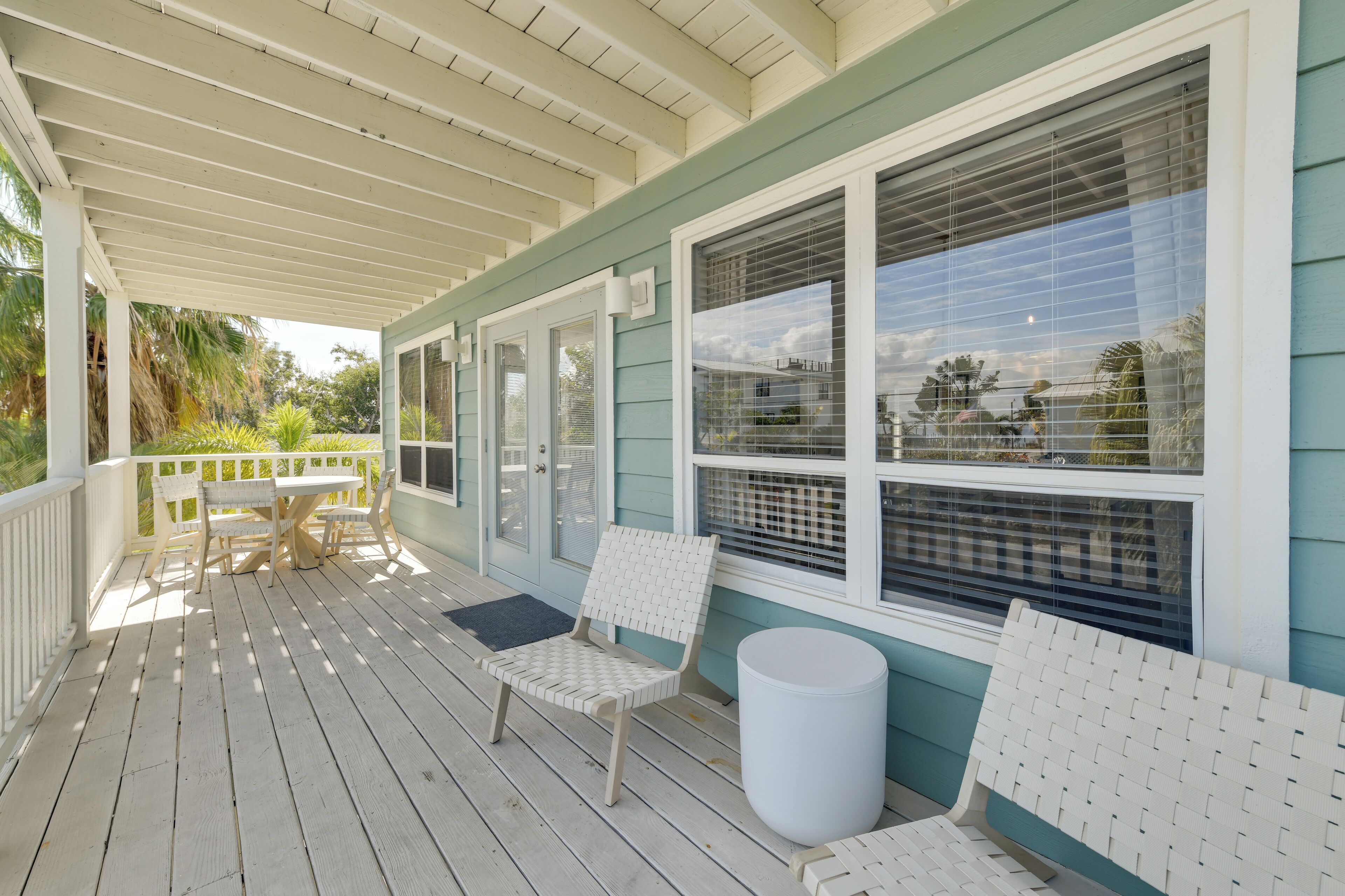 A covered porch with a table and chairs, a white trash can, and a window with blinds.