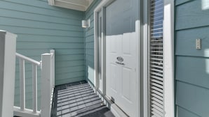 Un porche couvert avec une balustrade blanche, une porte avec une plaque nominative et une fenêtre avec des stores.