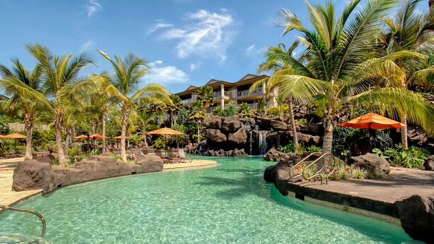 Ein klares Schwimmbecken mit Wasserfall, umgeben von Palmen und einem Resort-Gebäude.