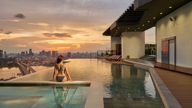 A woman sitting by an infinity pool with a city skyline in the background. - THE PENINSULA PHNOM PENH (Phnom Penh)