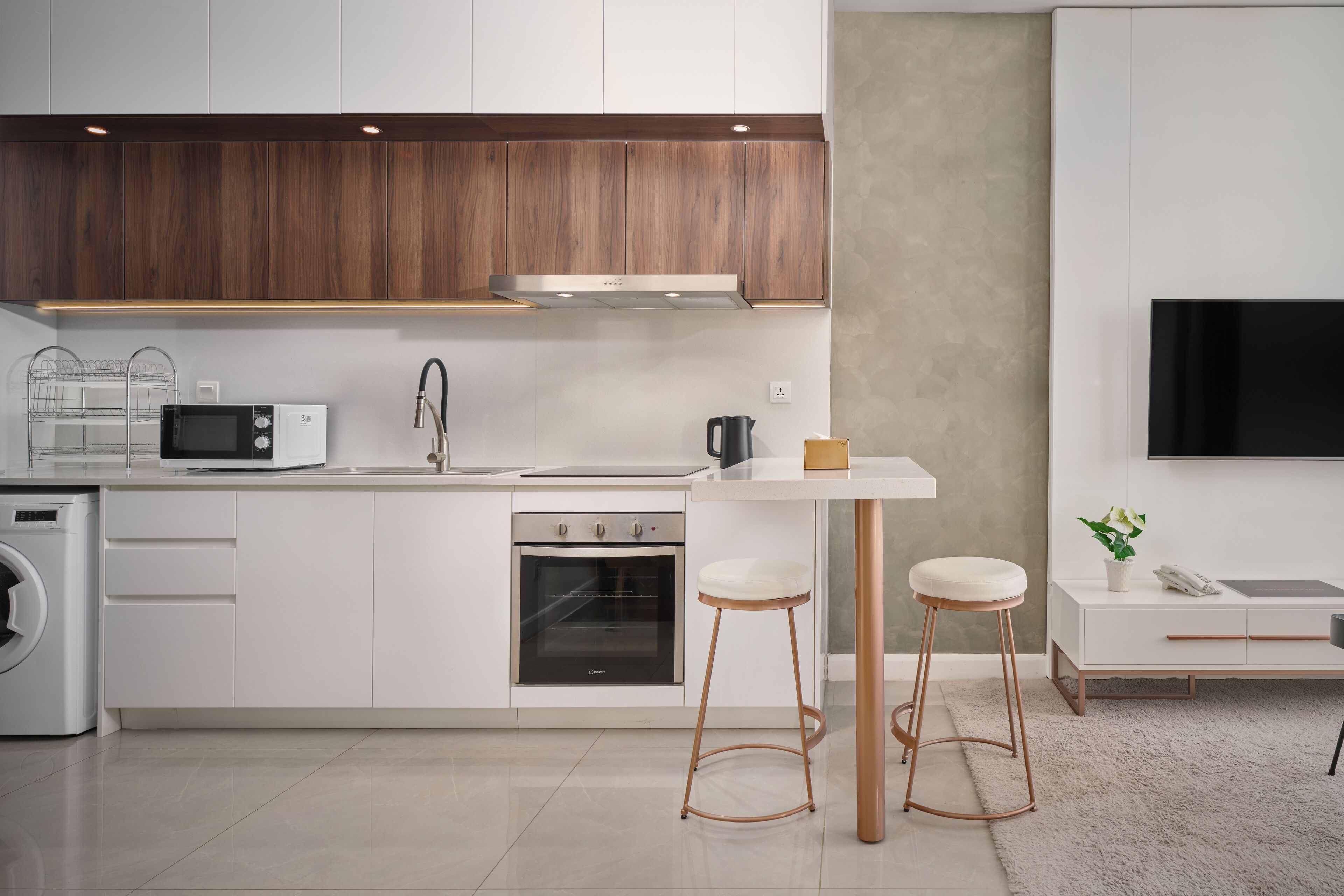Una cocina moderna con armarios blancos, horno integrado, microondas y cafetera. Hay dos taburetes y una mesita.
