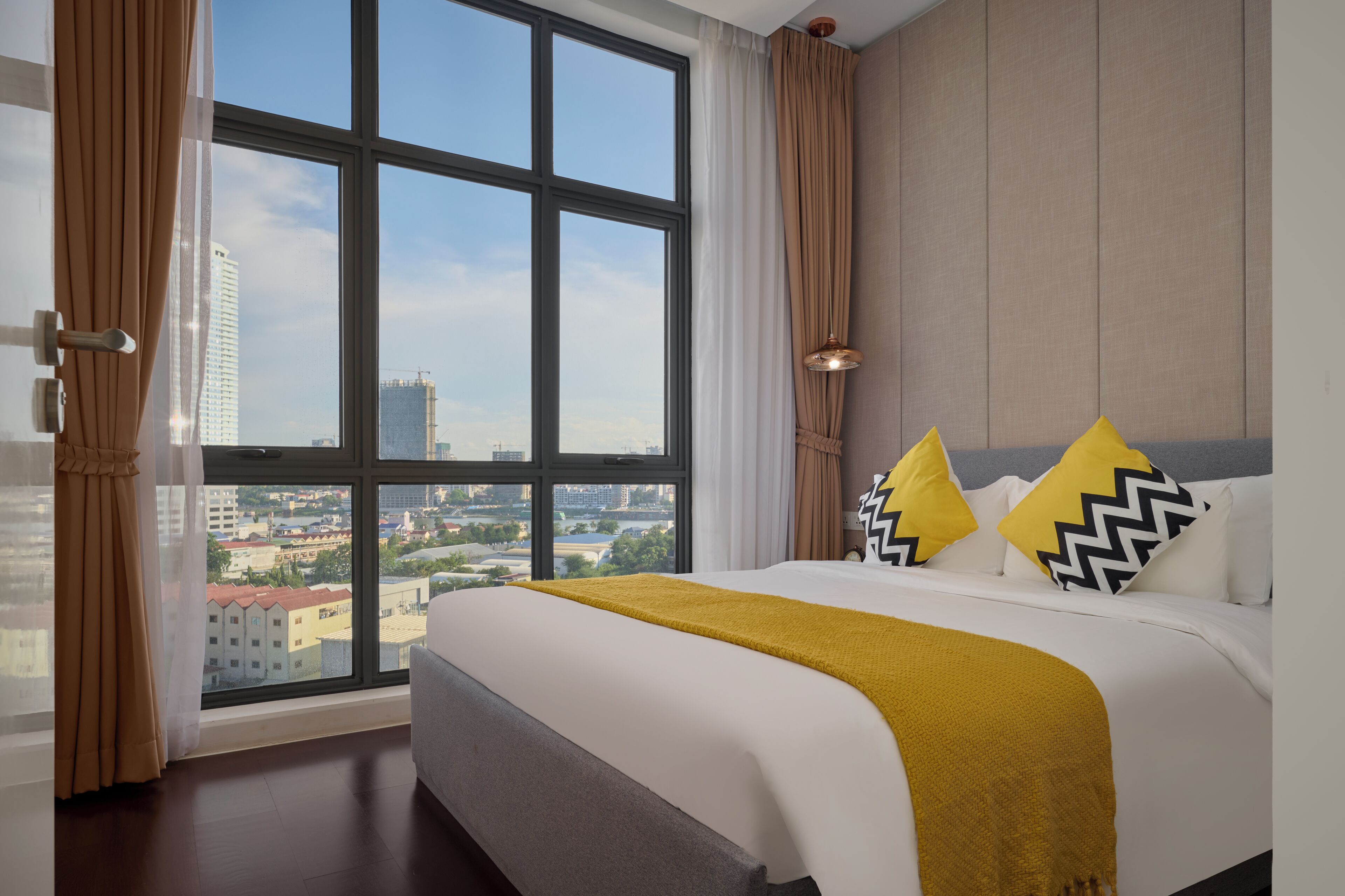A hotel room with a large window, a bed with white bedding and a mustard-colored throw, and two pillows with black and white patterns.