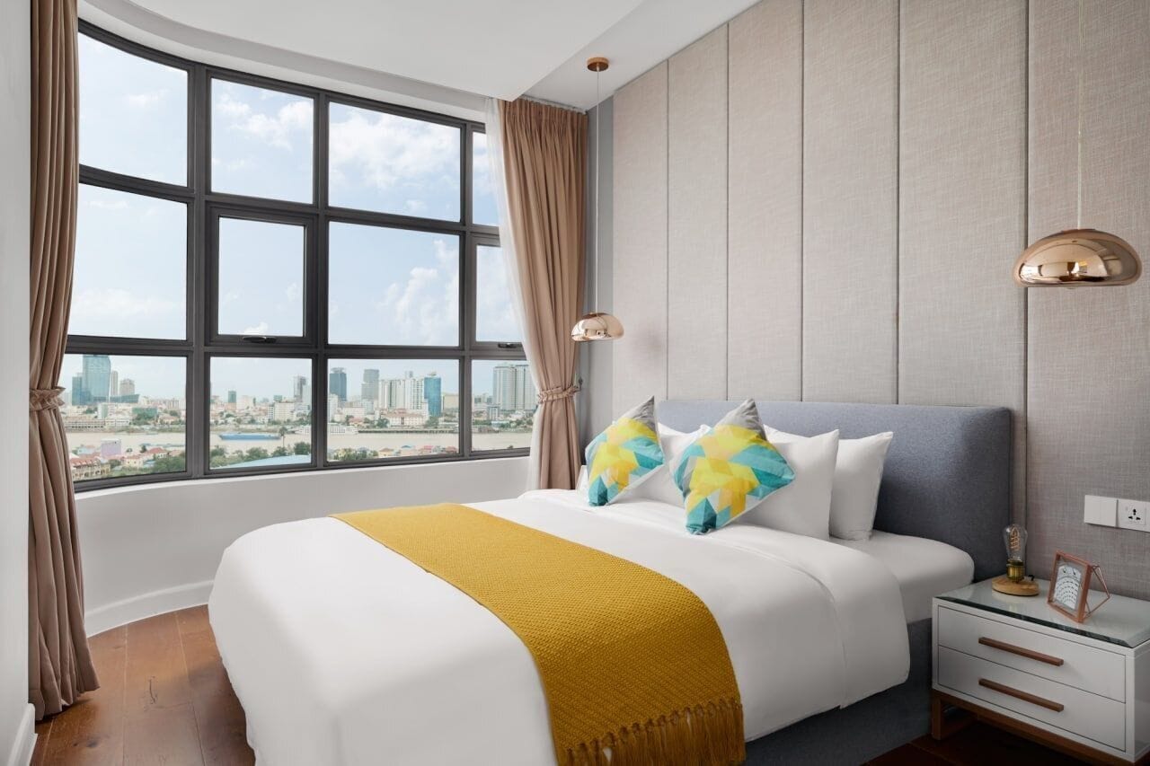 A modern hotel room with a large window offering a city view, a neatly made bed with a yellow throw, and a bedside table with a lamp and a picture frame.