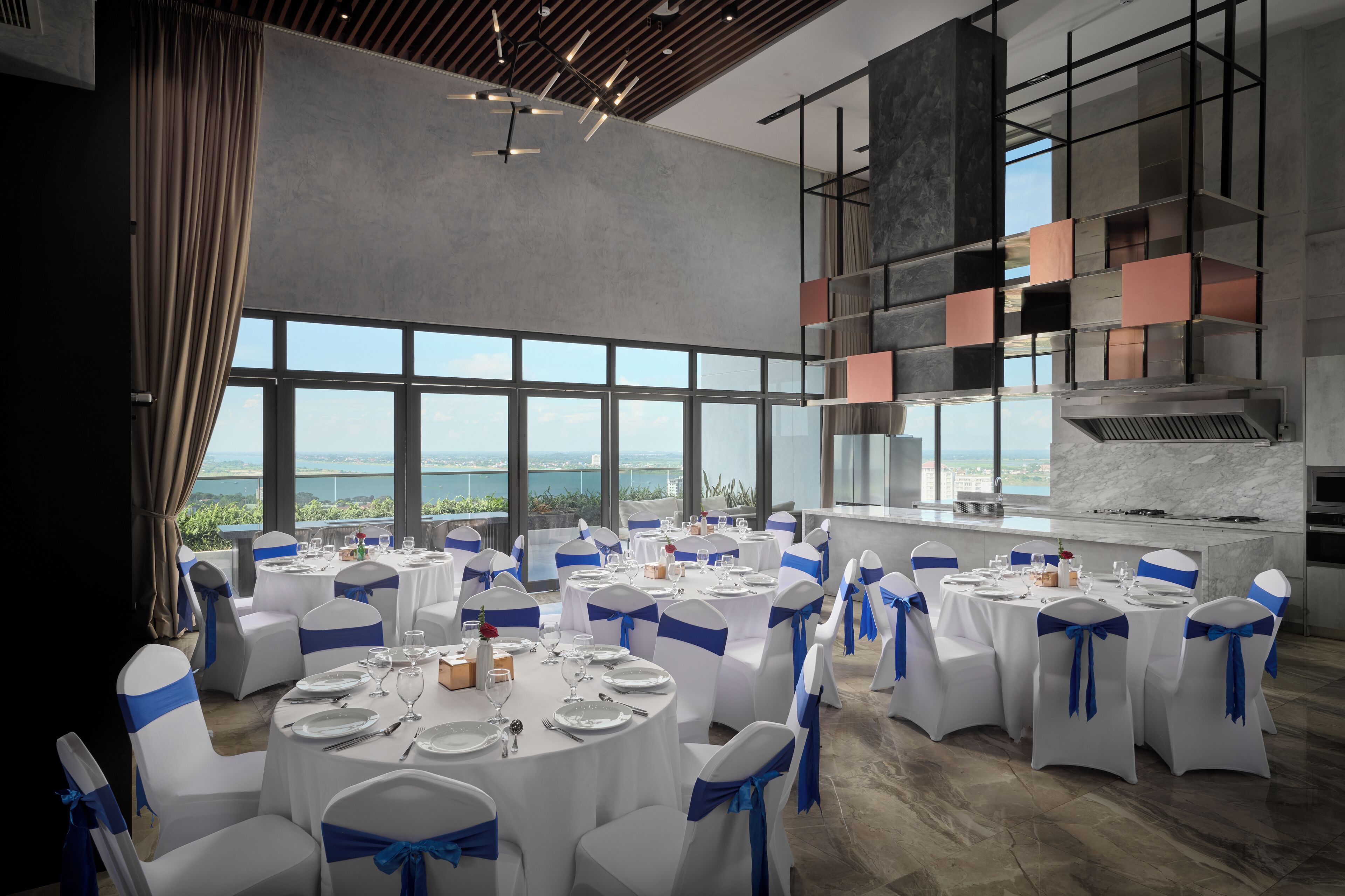 A spacious dining area with round tables and chairs, large windows offering a view of the sea, and a modern kitchen visible through the windows.