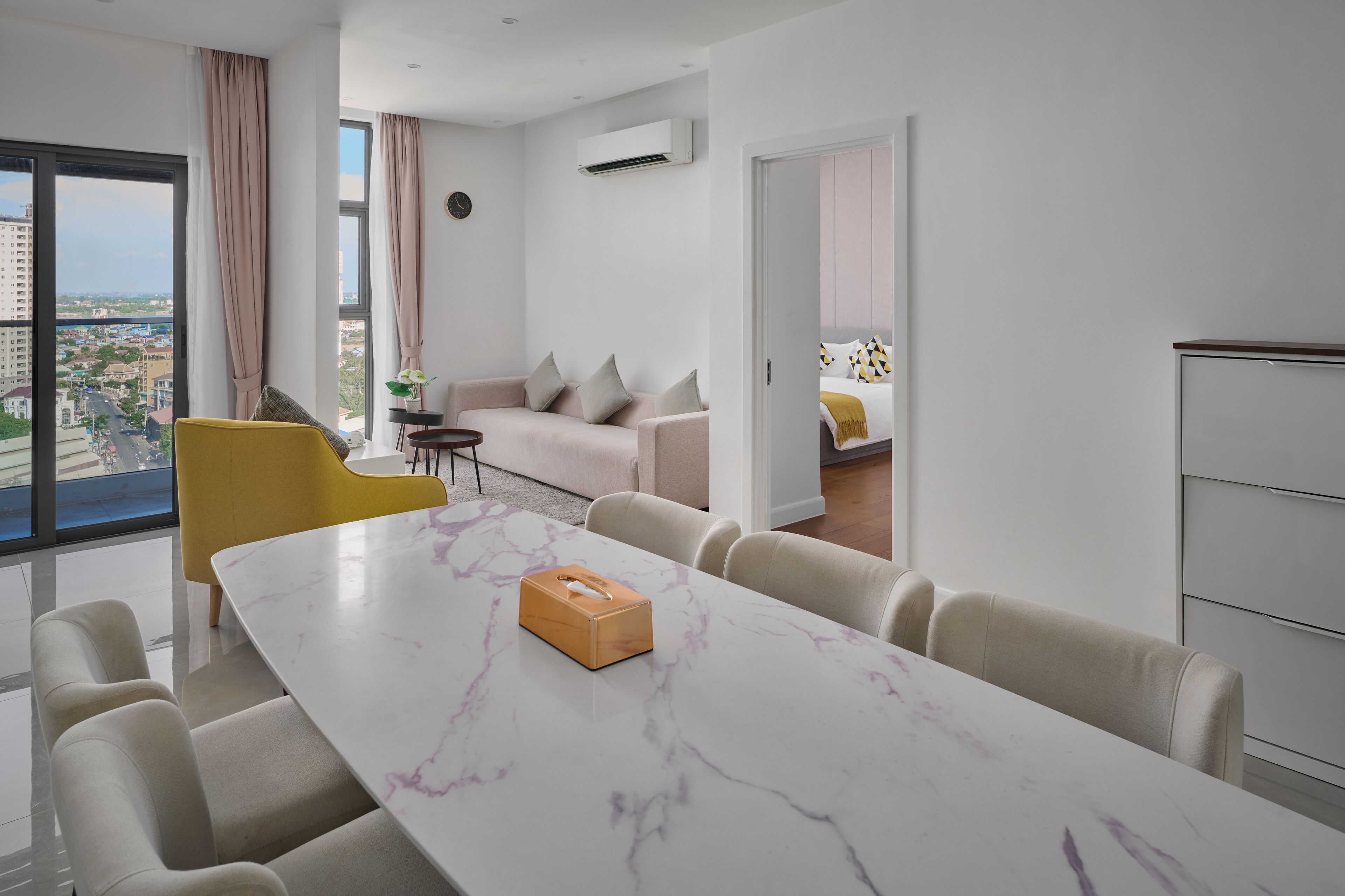 A modern hotel room with a marble dining table, a yellow chair, a sofa, and a view of the city through large windows.