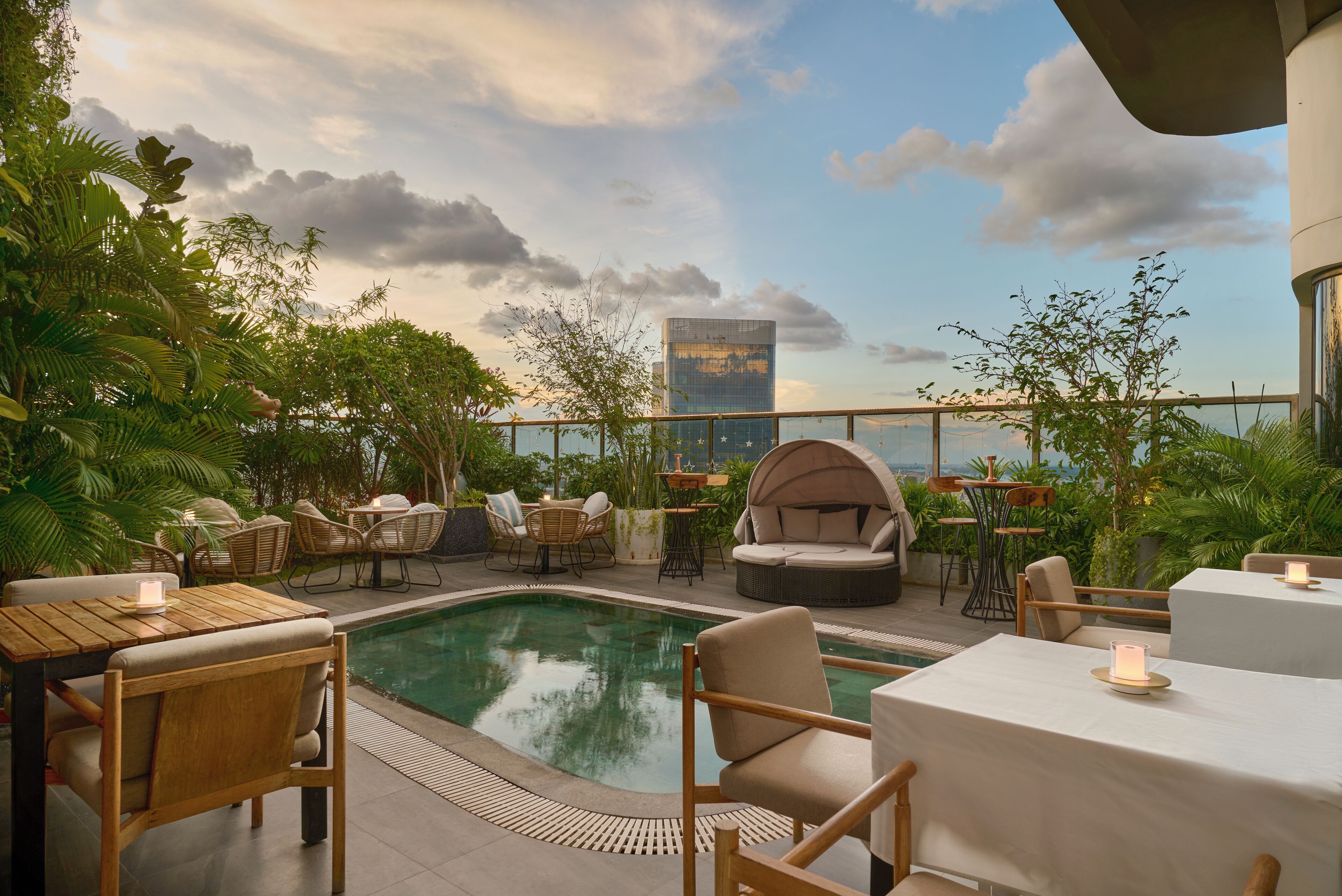 A rooftop pool area with seating and tables, surrounded by greenery and a cityscape view.