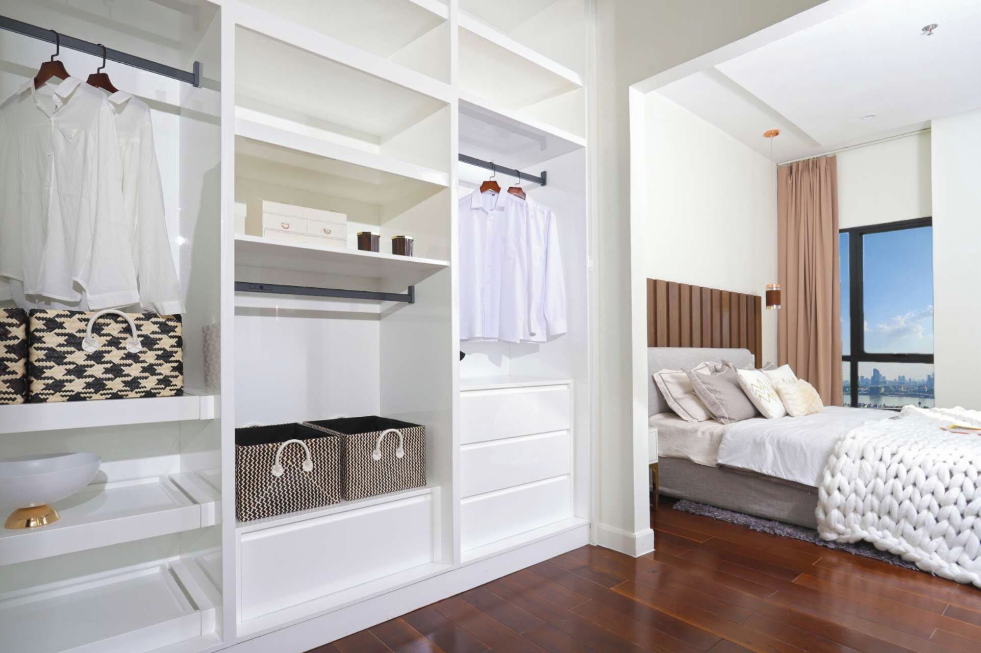 A modern bedroom with a large bed, a walk-in closet with hanging clothes and storage bins, and a city view through the window.