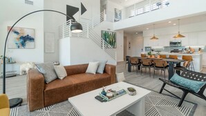 A spacious living area with a brown leather sofa, a white coffee table, and a patterned rug. A staircase leads to an upper level with a balcony.