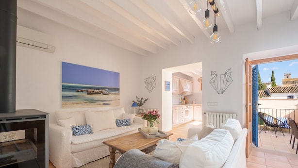 A modern living room with a white sofa, a wooden coffee table, and a painting of a beach scene. There is a kitchen visible through an open doorway.