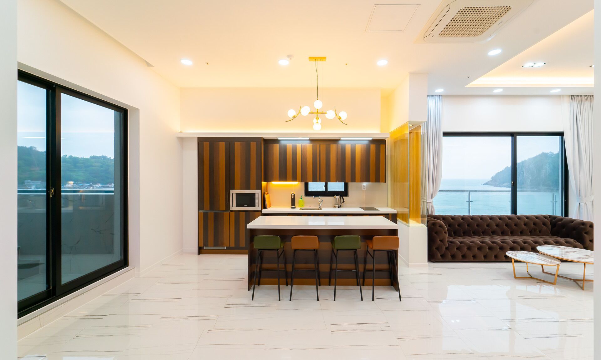 A modern kitchen with a central island, a dining area with a sofa and coffee table, and a large window offering a scenic view.