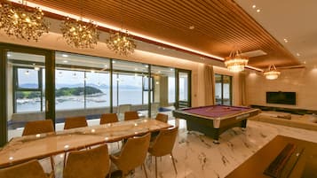 Una habitación moderna con mesa de billar, una mesa de comedor con sillas y ventanas amplias con vistas al mar.