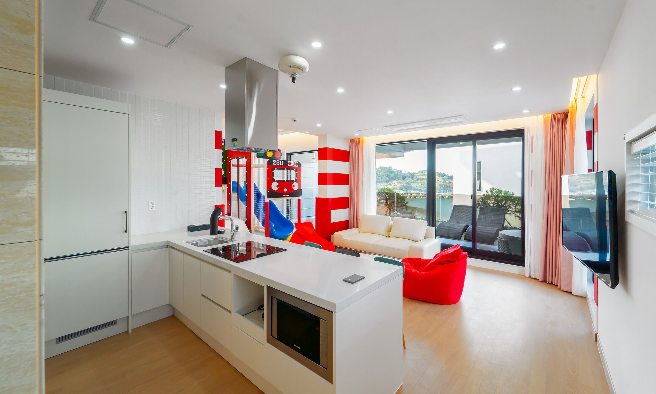 A modern kitchen with a central island, a dining area with a red bean bag, and a large sliding glass door leading to a balcony with a view.
