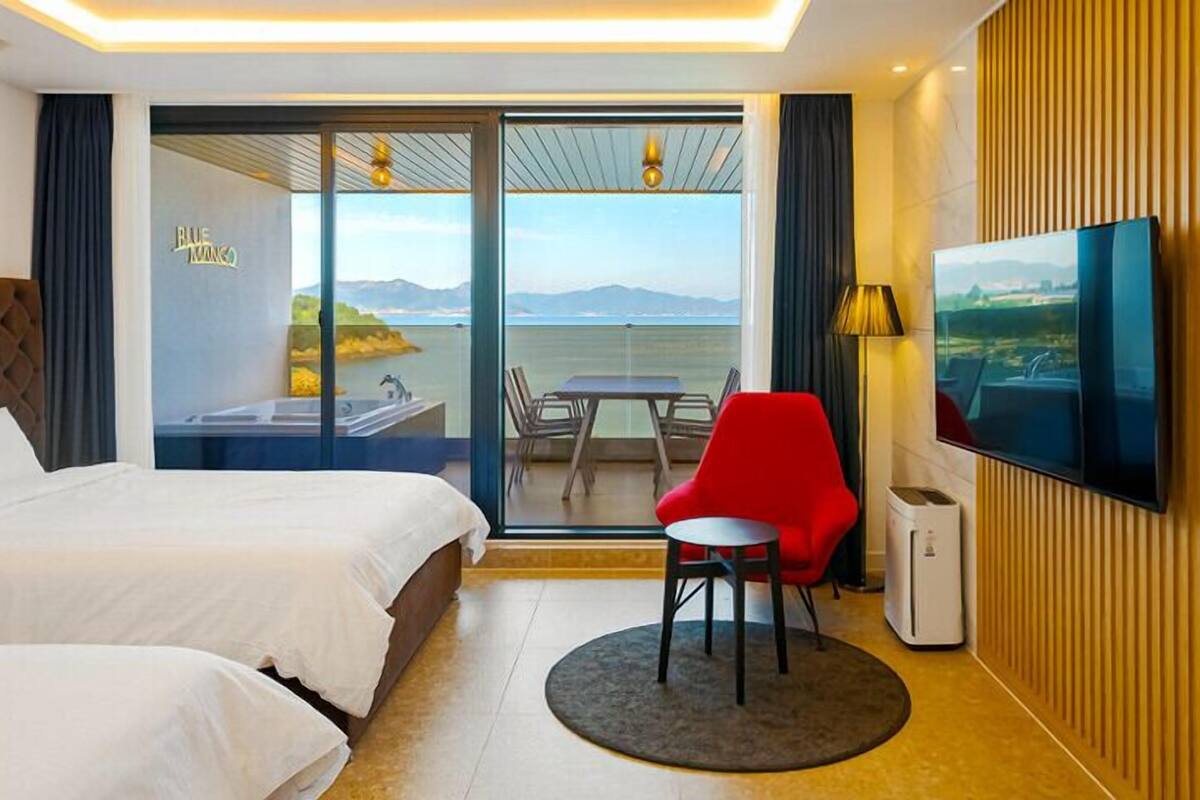 Une chambre d’hôtel avec un lit, une chaise rouge, une télévision et un balcon donnant sur la mer.