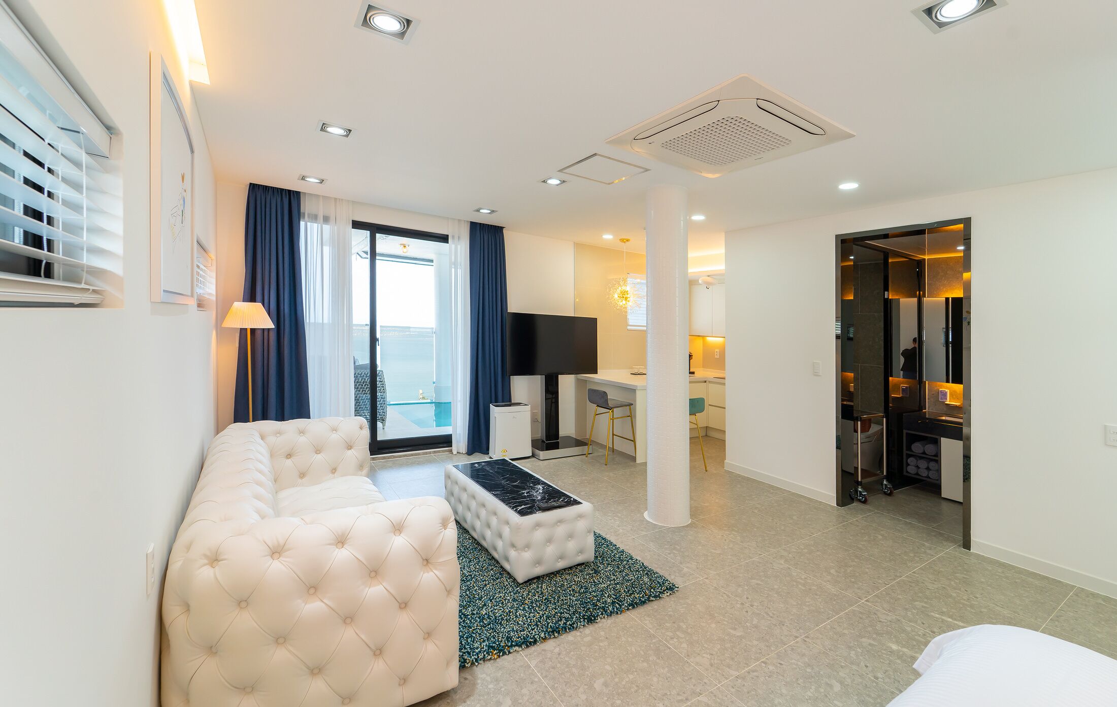 A modern living room with a sofa, a coffee table, and a TV. There is a balcony with a view, a desk, and a chair.