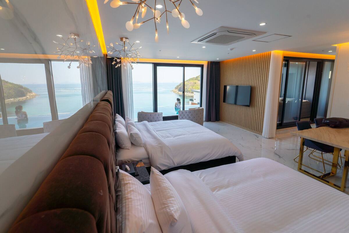 Une chambre d’hôtel moderne, dotée d’un grand lit, d’un canapé, d’une table à manger et d’un téléviseur. La chambre dispose de grandes fenêtres offrant une vue sur la mer.