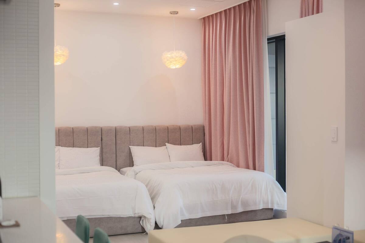 Une chambre d’hôtel avec deux lits, une grande fenêtre avec des rideaux roses et un luminaire moderne.