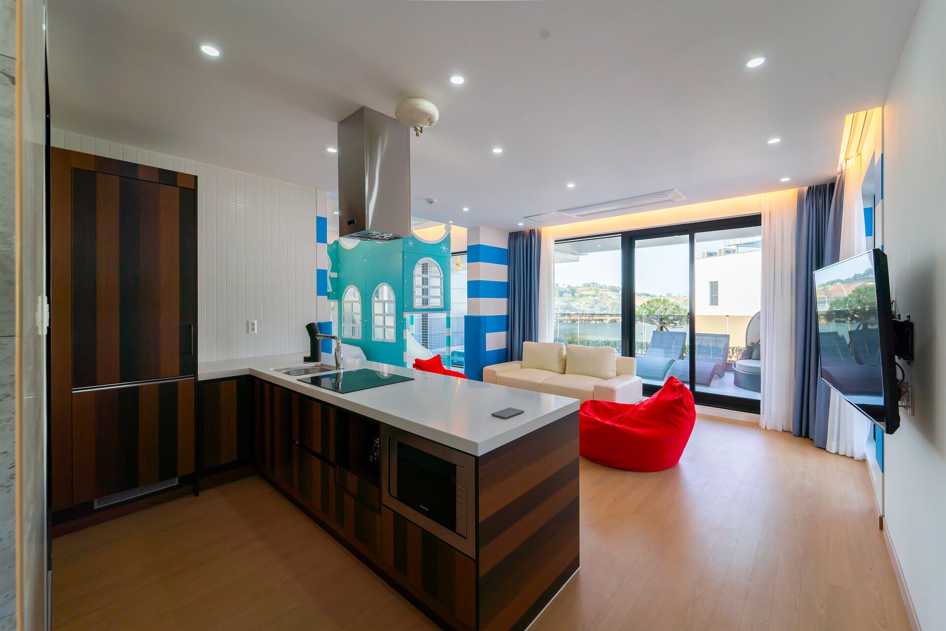 A modern kitchen with a central island, a dining area with a sofa and red bean bag, and a large window offering a view of the outdoors.
