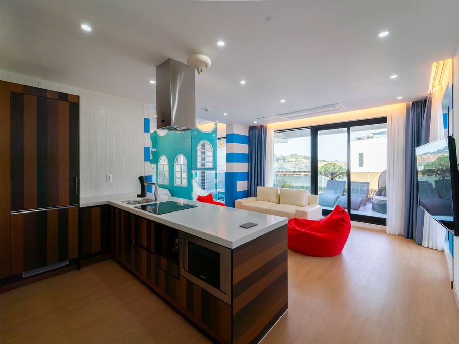 A modern kitchen with a central island, a dining area with a sofa and red bean bag, and a large window offering a view of the outdoors.
