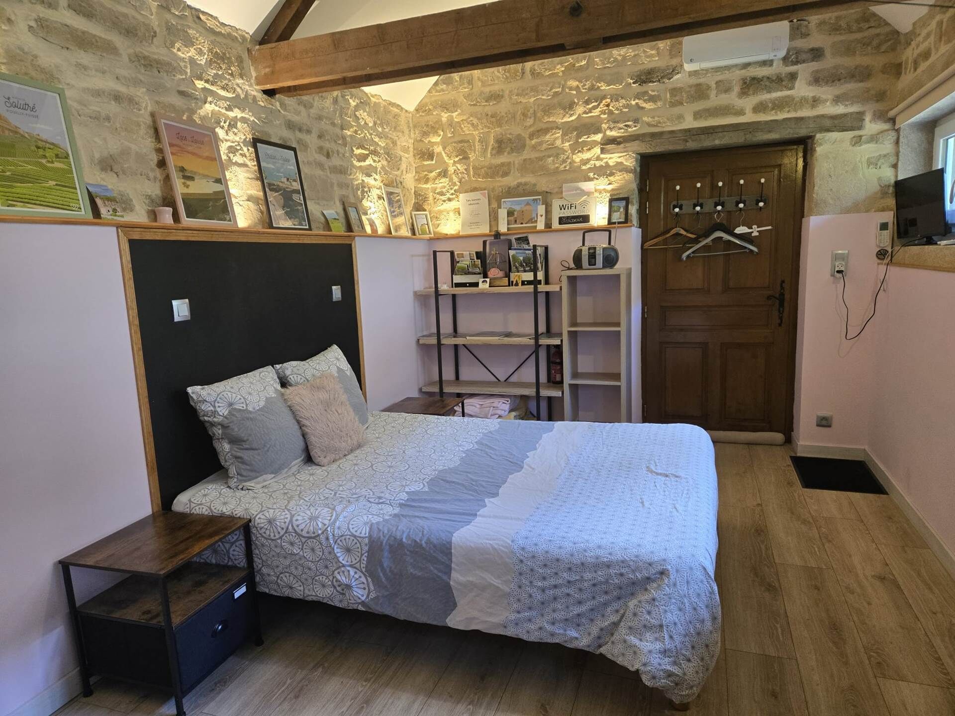 A bedroom with a bed, a wooden nightstand, a shelf with various items, a door, and a stone wall.