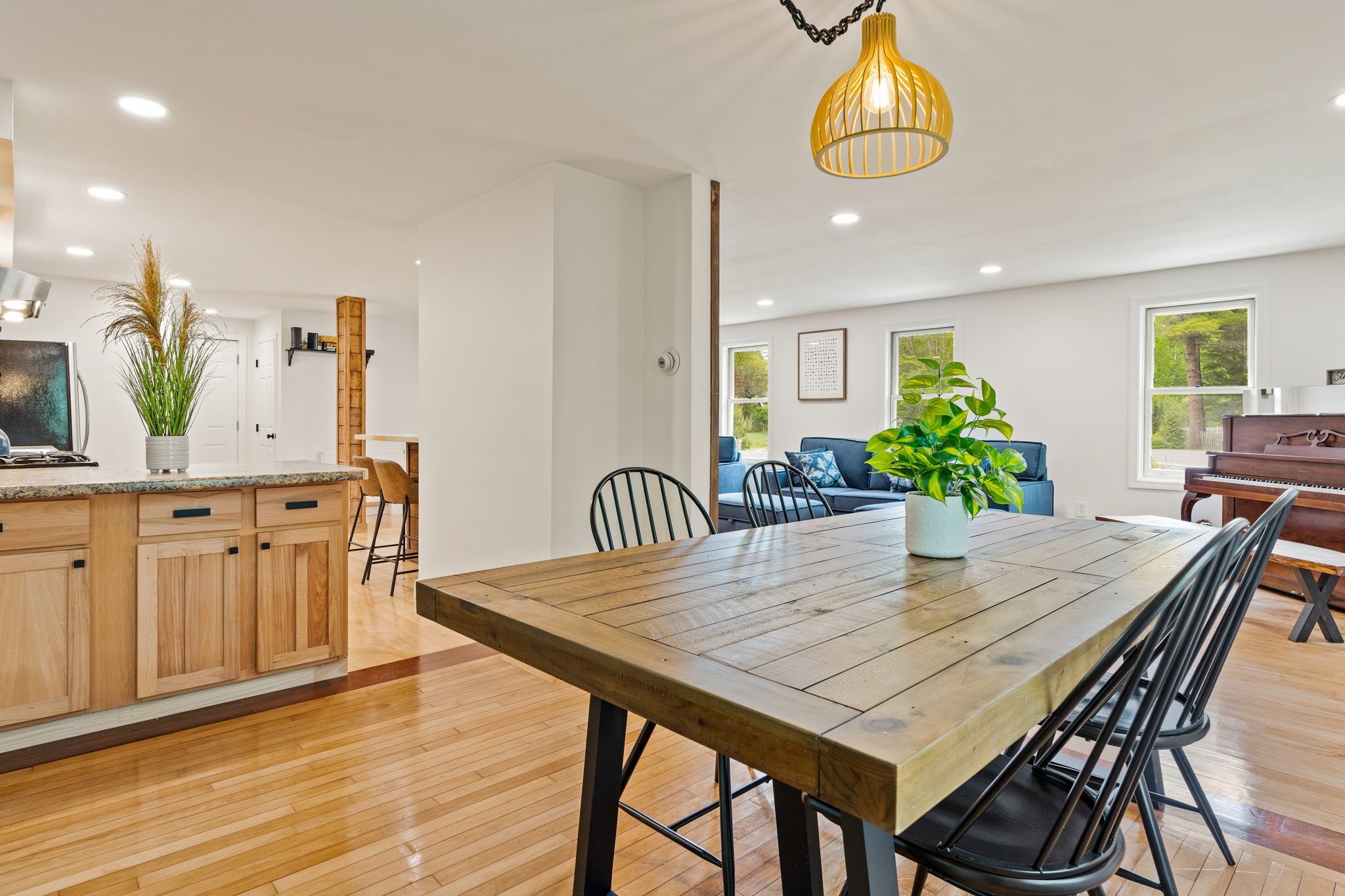 A modern kitchen with wooden cabinets, a dining table, and chairs. There is a large window with a view of greenery outside.
