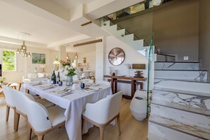 A modern dining area with a large table set for six, a staircase with glass railings, and a kitchen in the background. - Luxury and Modern villa with magnificent sea view & Gulf of Saint Tropez (Cavalaire-sur-Mer)