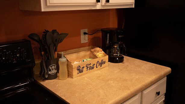 Una encimera de cocina con una cafetera, una caja con la etiqueta “But First Coffee” y un recipiente con utensilios.
