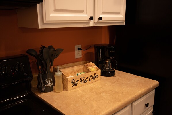 A kitchen counter with a coffee maker, a box labeled "But First Coffee," and a container of utensils.